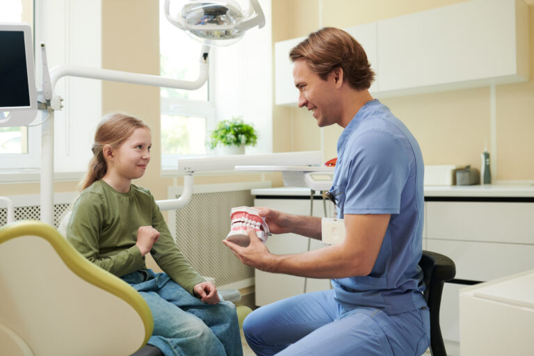 Caucasian male dentist showing dental model to Caucasian girl child sitting in dental chair, both smiling and interacting in modern dental clinic with equipment visible