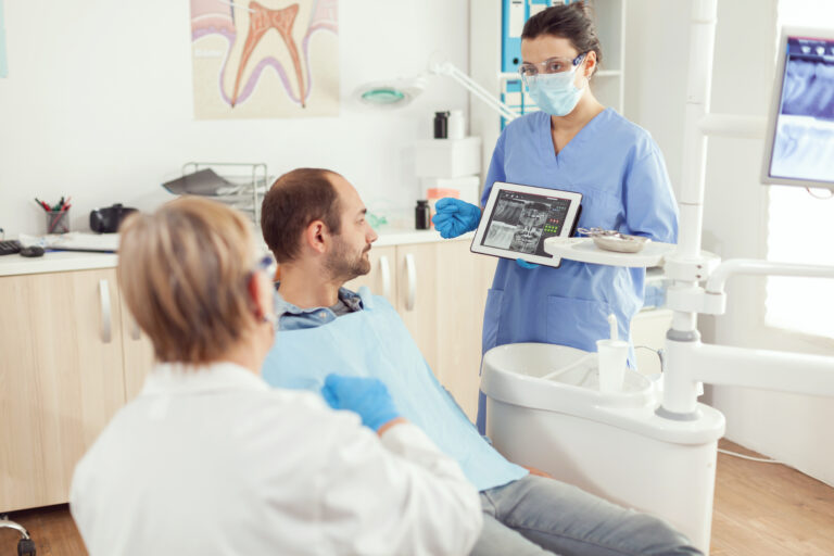 Stomatology nurse in dentistiry office showing tablet pc with tooth radiography explaining toothache treatment. Man sick patient sitting on dental chaiar in hospital orthodontist clinic office